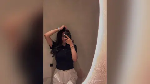 Young woman in a black polo shirt and white trousers taking a moody mirror selfie in a dimly lit room.
