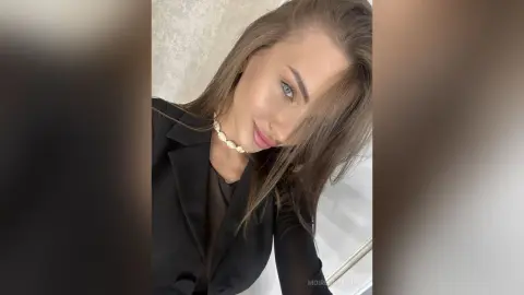 Elegant woman in a black blazer and gold necklace posing with a soft, glamorous expression indoors.