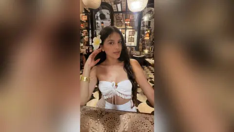 Young woman in a white boho crop top with a flower in her hair, posing in a cozy, dimly lit cafe.