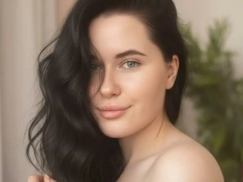 Close-up portrait of a smiling young woman with long wavy dark hair in a soft, natural indoor setting.