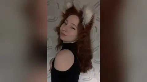 MadgeRemerez poses with a slight smile, wearing a black cut-out top and white furry ears against a patterned grey wall.