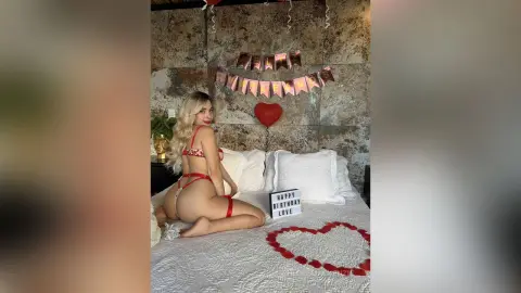 Blonde woman in red lace lingerie sitting on a white bed with heart decorations for a romantic birthday mood.