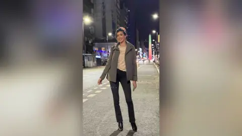 HaleyLyon stands on a city street at night wearing a grey jacket, a light tan top, and black skinny jeans.