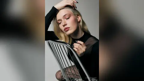 Elegant blonde woman in a black sheer top posing thoughtfully against a minimalist grey background.