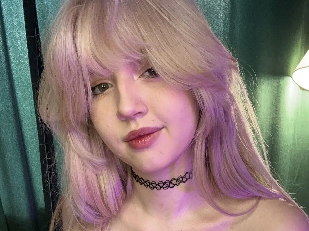 LucyStars poses with a slight smile, showing her long blonde hair and black lace choker against a teal curtain background.