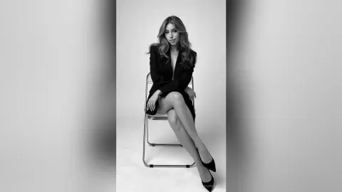 MeganLewit sits on a chair wearing a black blazer and black heels, posing with her legs crossed against a white background.