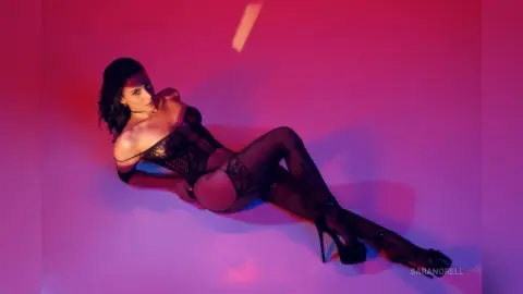 Elegant woman in black lace lingerie and high heels reclining against a moody pink studio background.