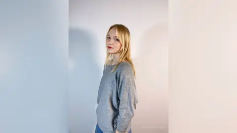Blonde woman in a grey long-sleeve sweater posing against a minimalist white background with soft lighting.