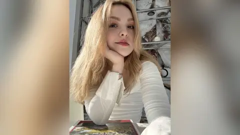 AliceTucci poses with her hand under her chin, wearing a white long-sleeved top with her long blonde hair down.
