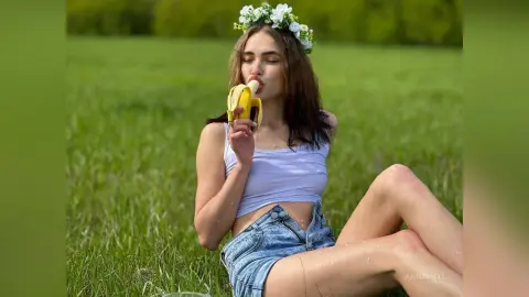 Young woman in white tank top and denim shorts eating a banana in a sunny green meadow.