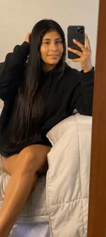 Kendra_Foox poses for a mirror selfie wearing a black hoodie, letting her long black hair hang down while sitting on a white bed.