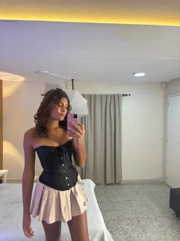 Young woman in a black corset and beige skirt taking a mirror selfie in a modern bedroom.