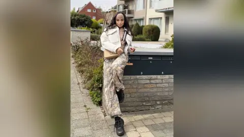 Stylish woman in camouflage pants and cream sweater leaning on a stone wall in an urban outdoor setting.