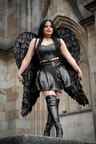 Woman in black leather outfit and large feathered wings posing regally against a stone cathedral background.