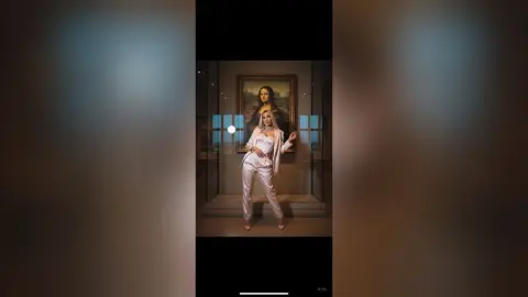 Elegant woman in a white jumpsuit posing in front of the Mona Lisa painting in a moody museum setting.