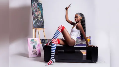 Young Black woman in a white bodysuit and striped socks posing playfully in an art studio with paintings.