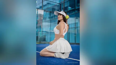 Young woman in white tennis outfit and visor sitting on a blue court against a modern glass building background.