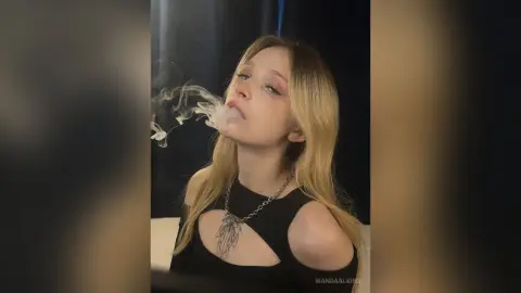 Blonde woman in a black cutout top exhaling smoke in a moody, dark studio setting.