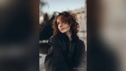 Red-haired woman in a black winter jacket looking away in a moody urban outdoor setting.