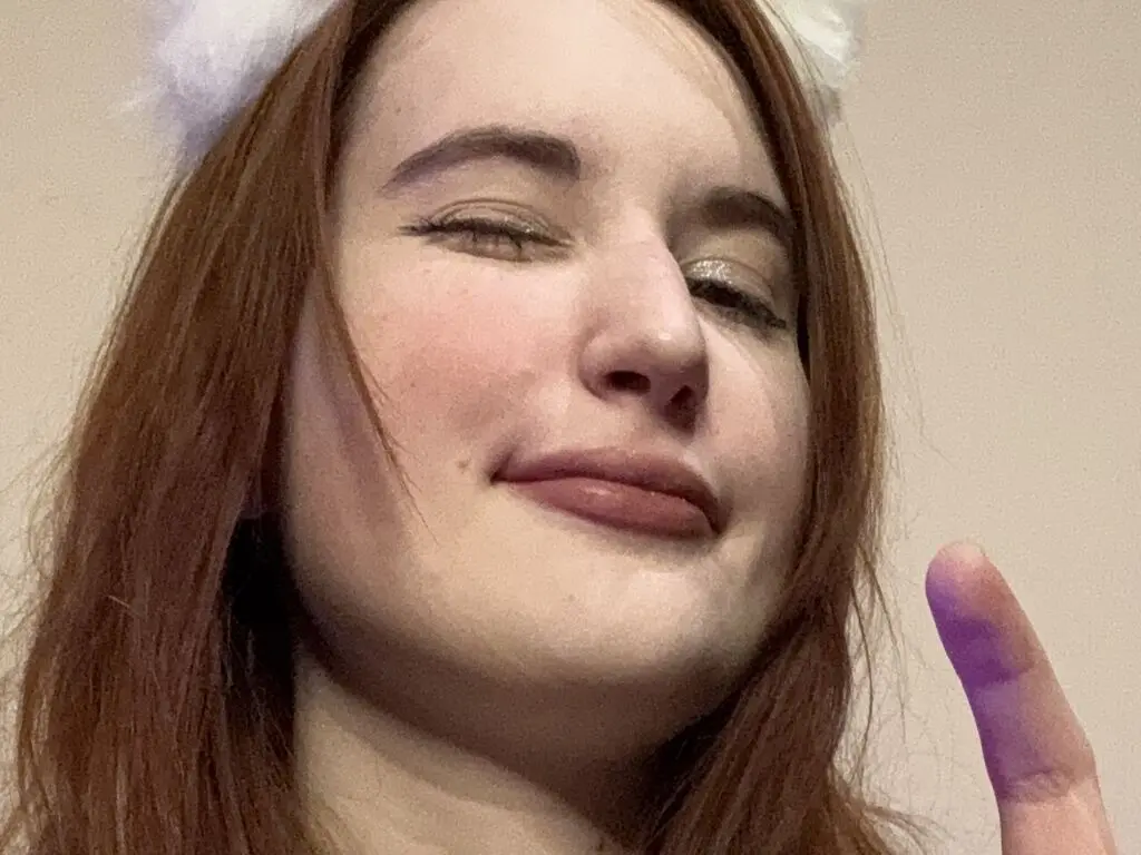 Smiling redhead woman with glitter eye makeup and white fuzzy headband in a cozy indoor setting.