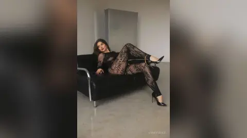 Elegant woman in a black lace bodysuit lounging on a modern black sofa in a minimalist studio setting.