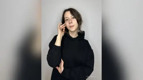 Young woman in a black oversized hoodie posing against a minimalist grey background with a calm expression.
