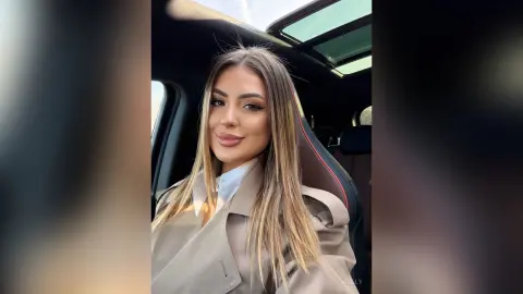 Smiling woman in a beige trench coat sitting in a car with a sunroof on a bright day.