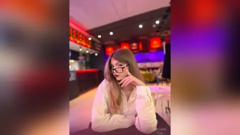 Young woman in white shirt and glasses sitting in a moody, dimly lit cafe with red ambient lighting.