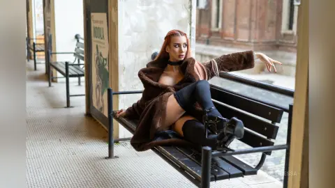 DeviNessa sits on a black park bench wearing a brown fur coat and black thigh-high stockings.