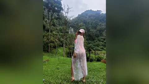 Young woman in a pink swimsuit and sheer white lace cover-up standing in a lush green tropical forest.