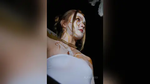 AliceTucci poses in a white top with gold liquid dripping down her face, neck, and chest against a dark background.