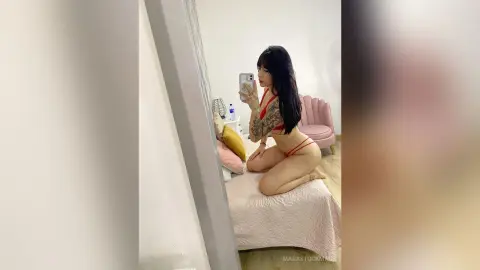 Young woman in red lingerie taking a mirror selfie while kneeling on a bed in a bright bedroom.