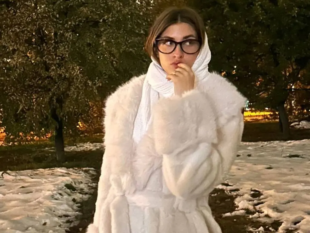 Stylish woman in a white faux fur coat and scarf posing in a snowy park at night.