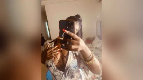 Young woman in a floral print top taking a mirror selfie in a cozy bedroom setting.