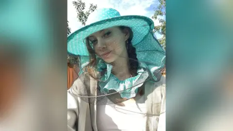 Smiling woman in a turquoise wide-brimmed sun hat and white outfit outdoors in bright sunlight.