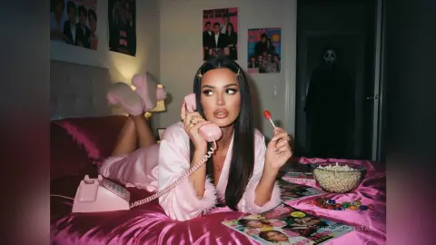 Woman in pink silk pajamas lying on a bed with a vintage pink telephone and lollipop in a nostalgic bedroom.