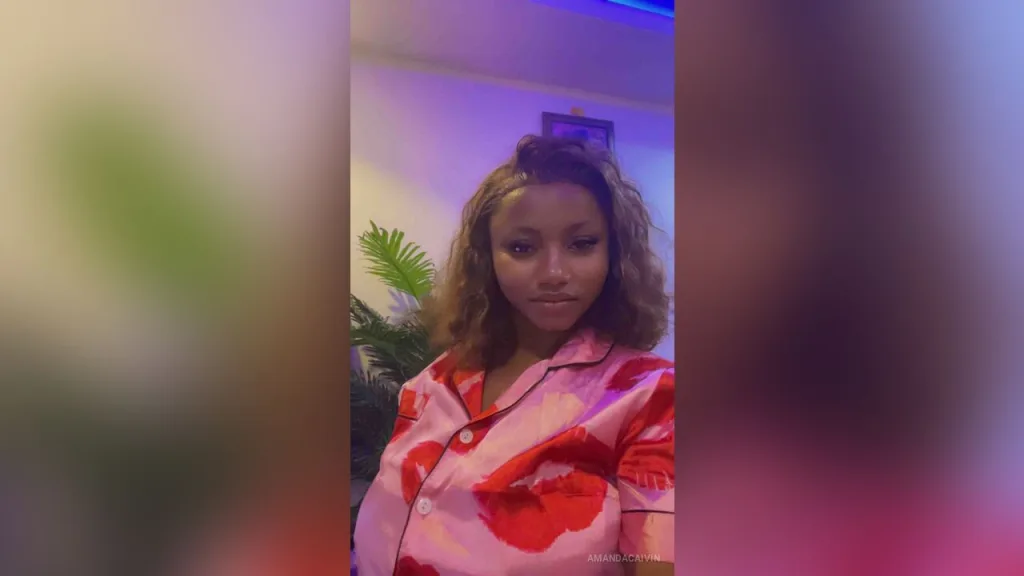 AmandaCaIvin — AmandaCaIvin wears pink and red patterned pajamas while looking at the camera in a room with plants.
