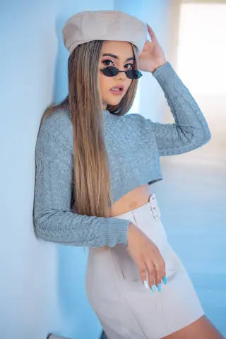 Young woman in a grey cable knit sweater, beige beret, and white skirt posing against a blue wall.