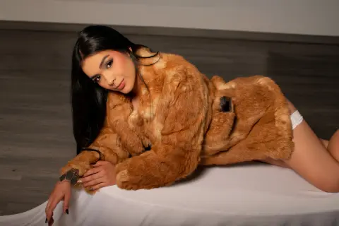 Young woman in a cozy brown faux fur coat lounging on a white surface with a relaxed, moody vibe.