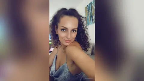 AishaApril poses in a low-cut silver top, showing her cleavage and curly dark hair while looking at the camera.