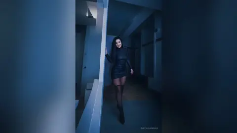 Smiling woman in a black long-sleeve mini dress and tights standing in a moody, dimly lit blue hallway.