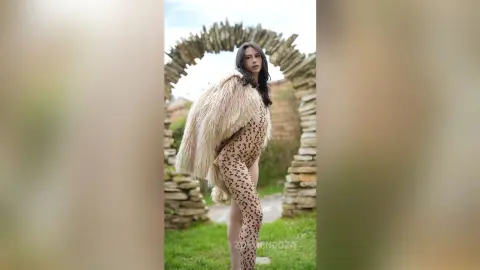 Elegant woman in a leopard print bodysuit and faux fur coat posing by a stone archway in a lush garden.