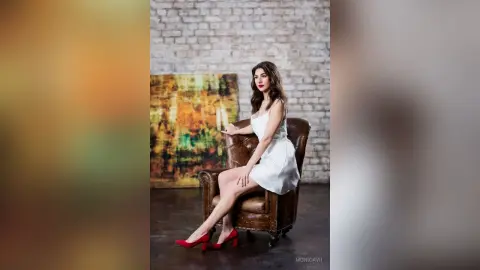 Elegant woman in a white cocktail dress and red heels sitting on a leather chair in a rustic loft setting.