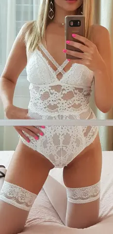 Woman in white lace bodysuit and matching lace thigh-high stockings posing in a bright bedroom.