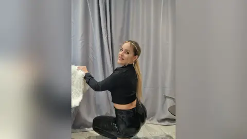 Smiling blonde woman in a black long-sleeve crop top and shiny black leggings posing in a studio setting.