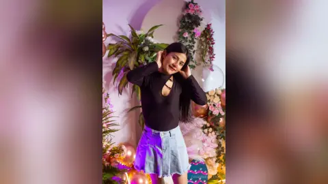Young woman in a black long-sleeve top and denim skirt posing happily in a floral decorated room.