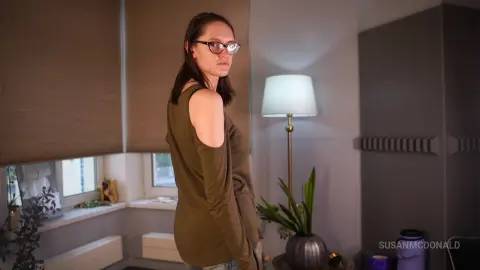 Thoughtful young woman in glasses and olive top standing in a dimly lit modern living room.
