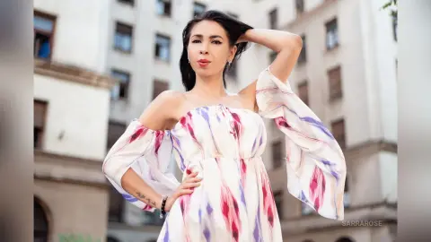 Stylish woman in a white and purple floral off-the-shoulder maxi dress posing outdoors in a city setting.