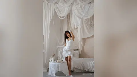 HermaCovel poses in a white lace teddy, running her hand through her long dark hair in a white draped room.