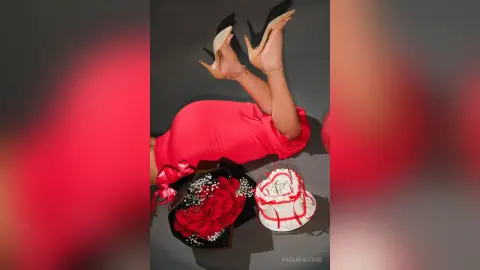Woman in a red dress lying on a gray floor next to a red rose bouquet and a white frosted cake.